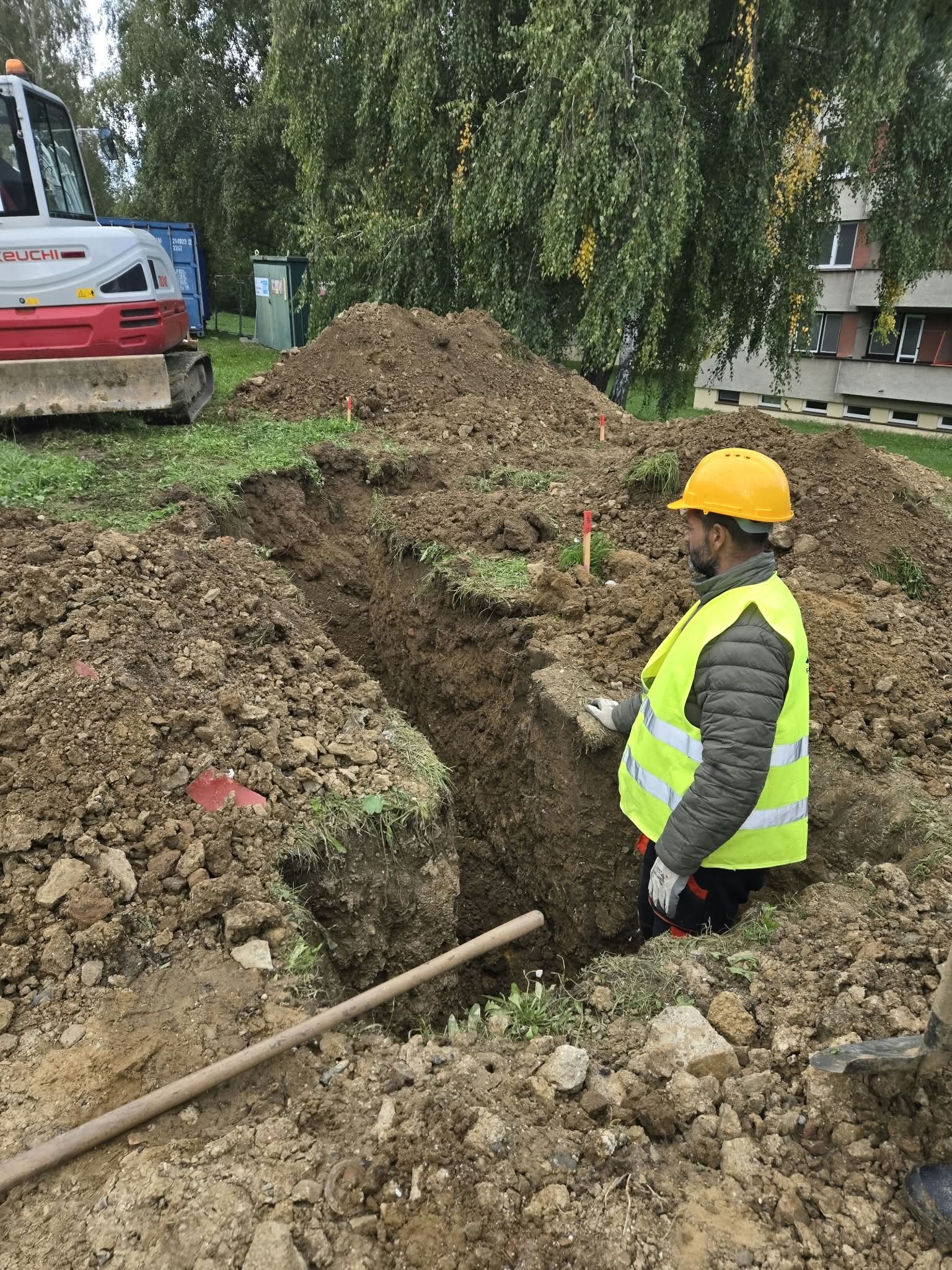 Robotník v prilbe ručne dočisťuje ryhu, za ním rýpadlo