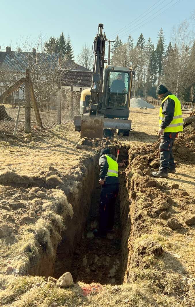 Robotník v hlbokej ryhe, nad ním rýpadlo a kolega v reflexnej veste