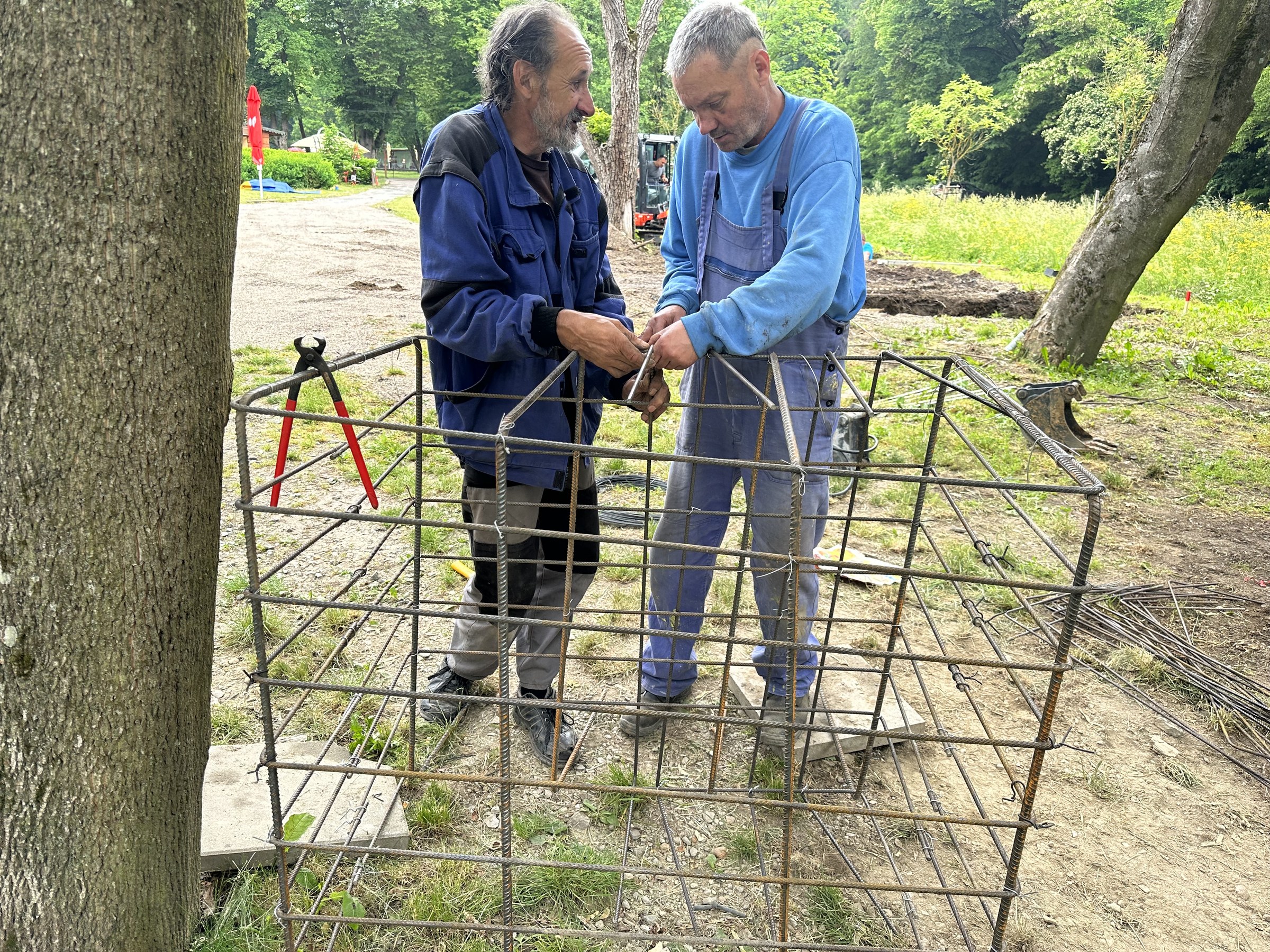 Dvaja robotníci viažu kôš výstuže pod stromami